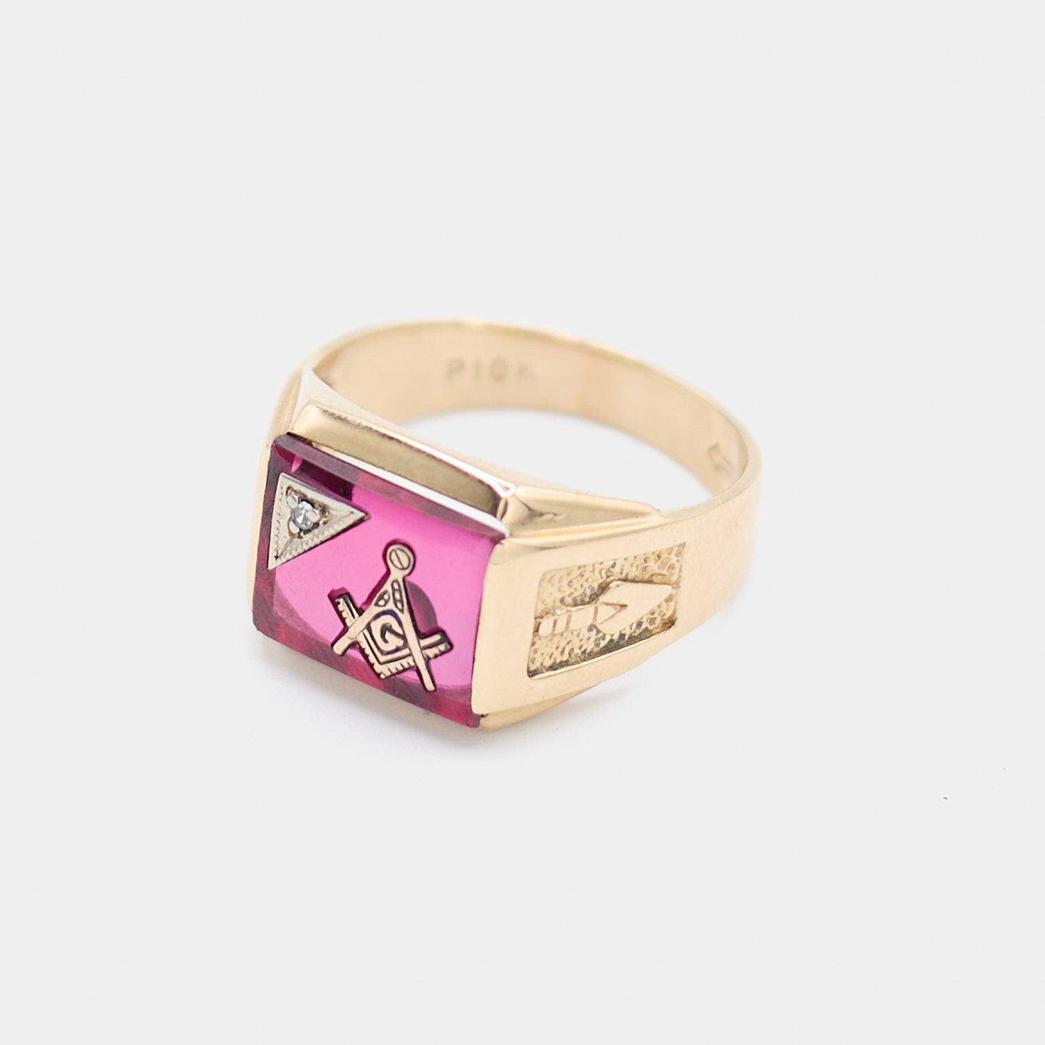 Close-up angled view of a pre-owned 10K yellow gold Freemason ring showing the Masonic square and compass design set in a red stone, with a small white stone accent and the 10K gold hallmark stamped inside the band.