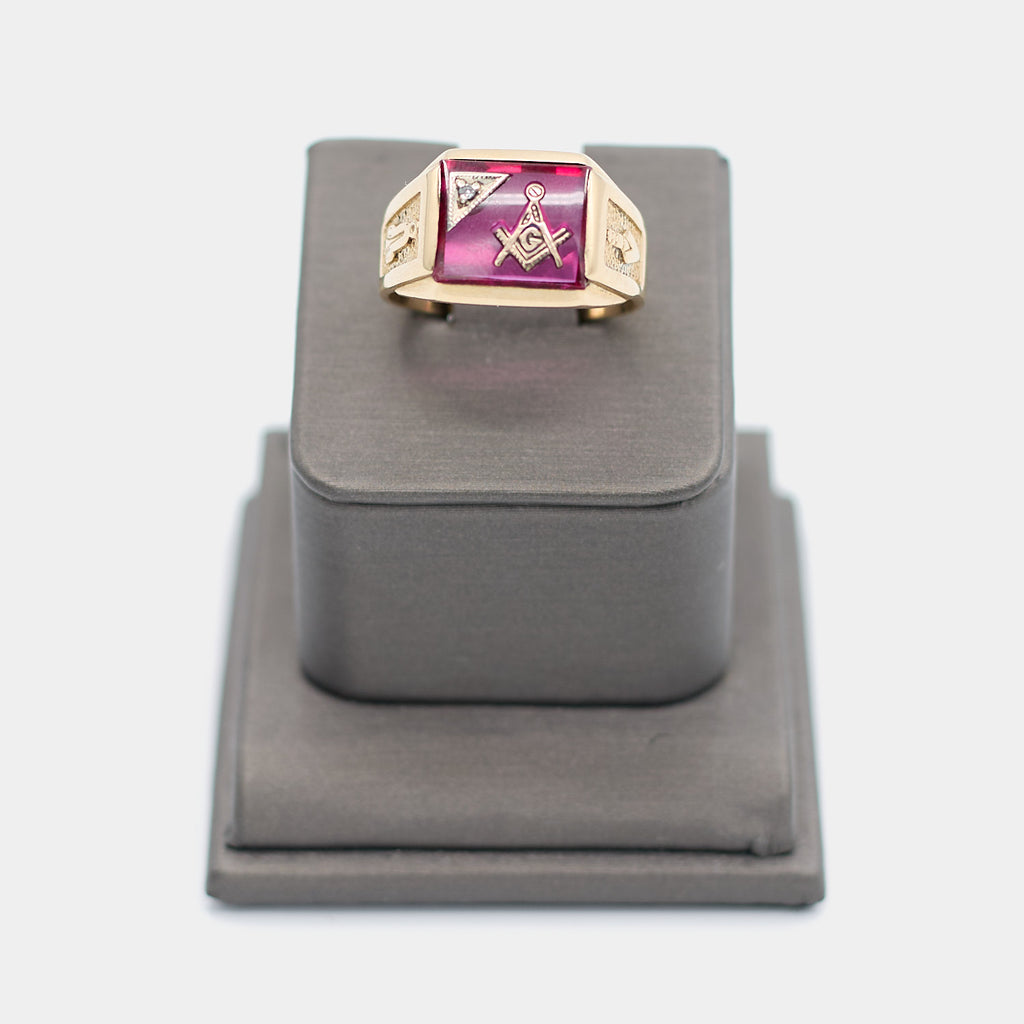 Pre-owned 10K yellow gold Freemason ring, size 9.5, featuring a large rectangular red stone with the Masonic square and compass emblem and a small white stone accent, shown front-facing on a display stand. Weighs 6.78 grams.