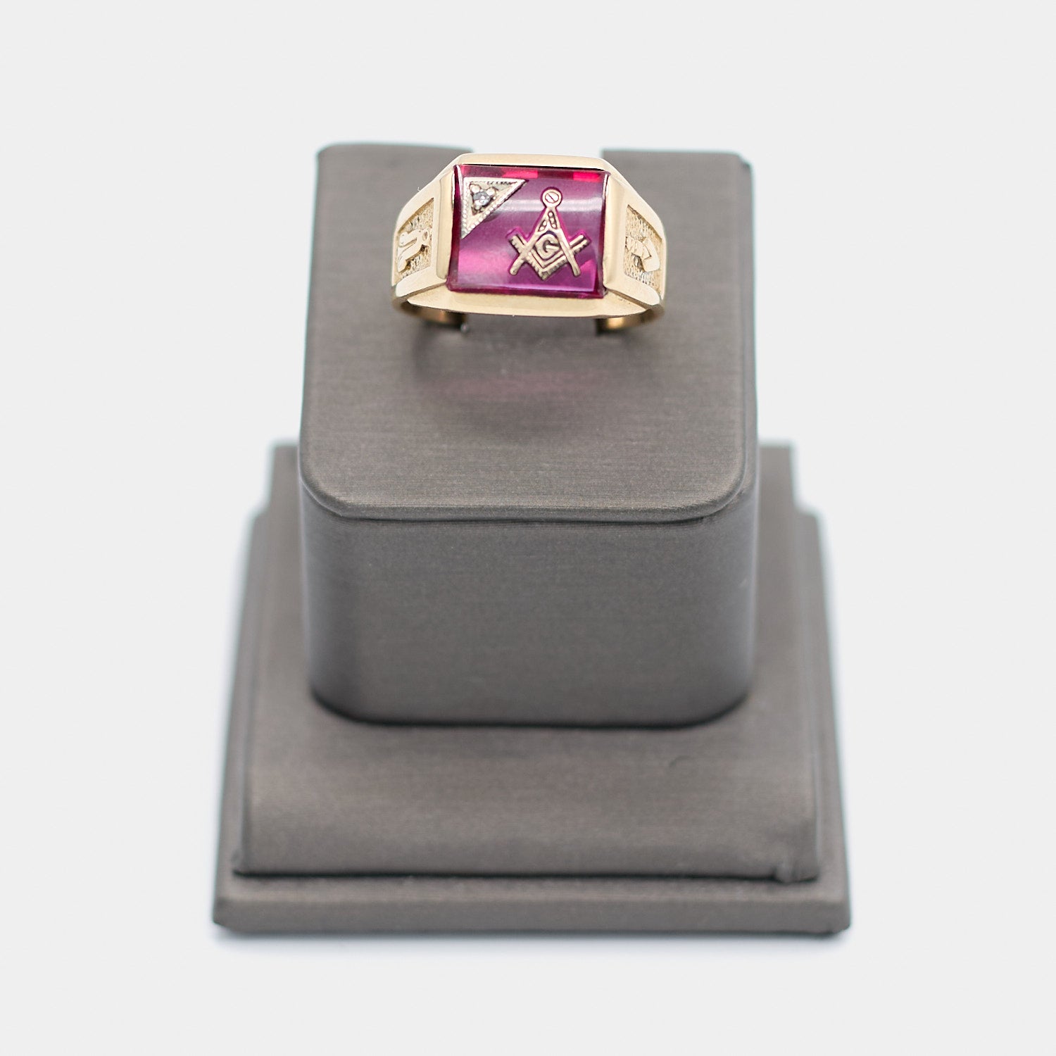 Pre-owned 10K yellow gold Freemason ring, size 9.5, featuring a large rectangular red stone with the Masonic square and compass emblem and a small white stone accent, shown front-facing on a display stand. Weighs 6.78 grams.