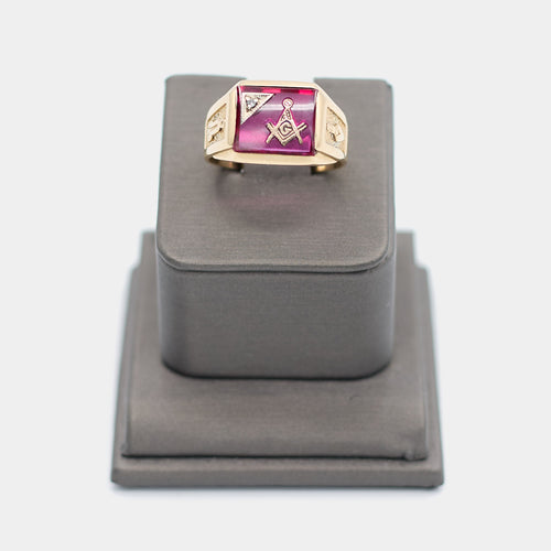Pre-owned 10K yellow gold Freemason ring, size 9.5, featuring a large rectangular red stone with the Masonic square and compass emblem and a small white stone accent, shown front-facing on a display stand. Weighs 6.78 grams.