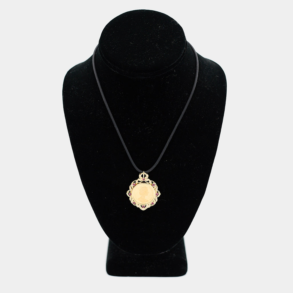 Pre-owned 18K yellow gold coin pendant featuring a 24K gold Isle of Man coin, set with red stones and diamonds, shown on a black cord necklace against a display bust.