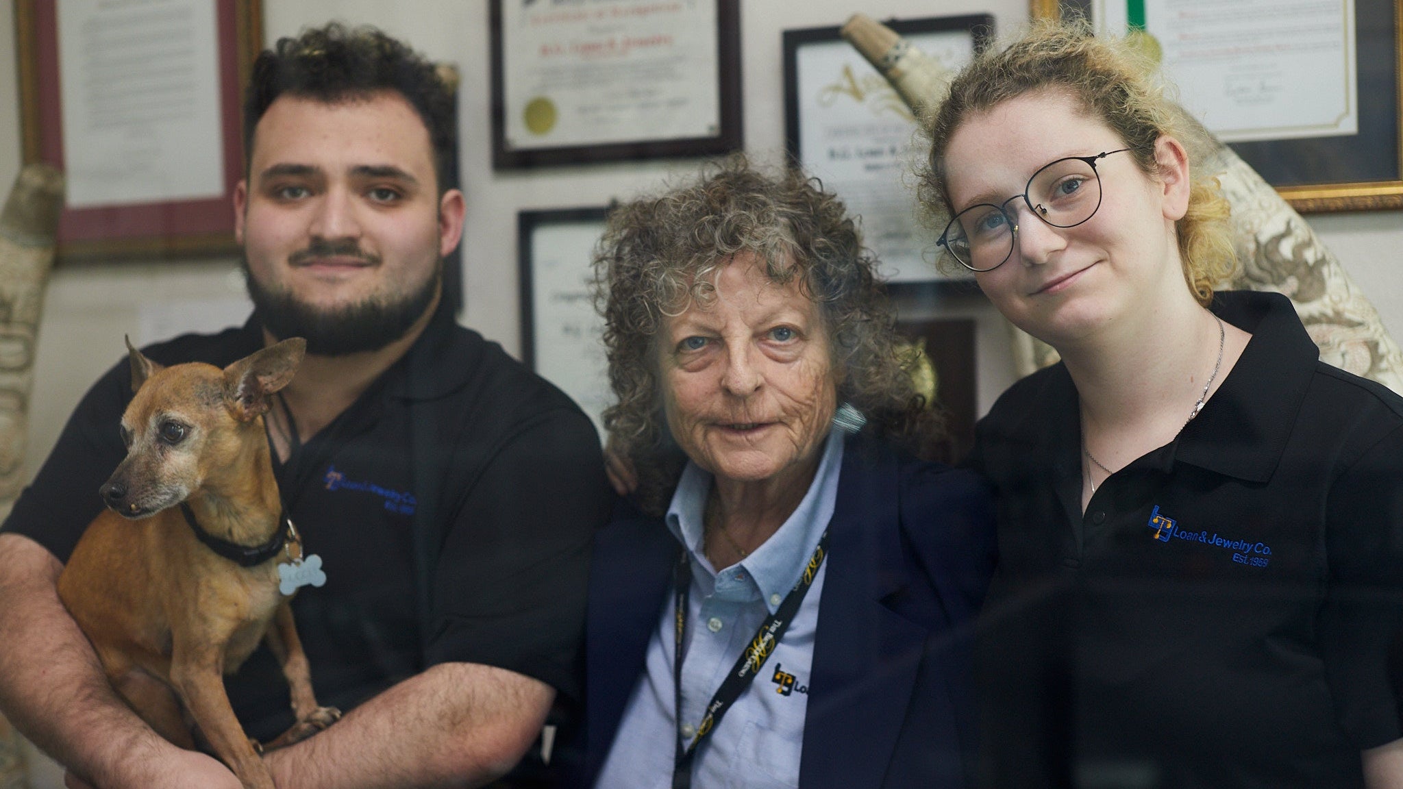 Three people with a small dog in an indoor setting with framed documents on the wall.